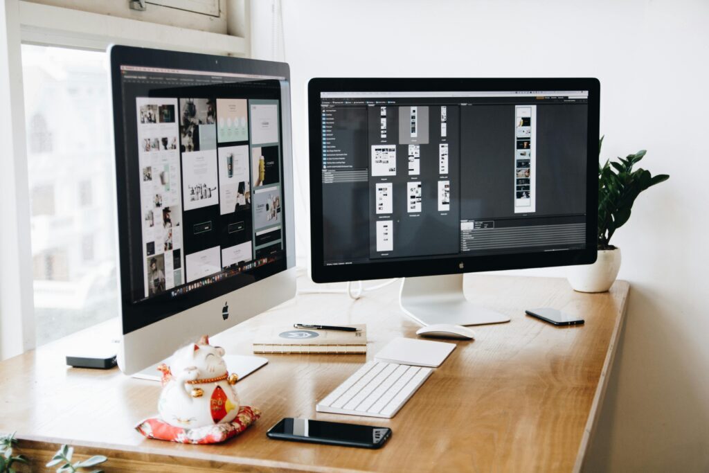 A modern workspace featuring two Apple desktop monitors displaying Website Design and Development software, with a keyboard, mouse, notebook, smartphone, lucky cat figurine, and a potted plant on a wooden desk near a window.